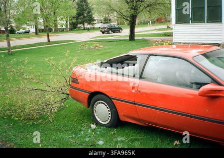 A red coupe with a crushed roof and broken window caused by a fallen tree branch in a residential neighborhood after a storm. Stock Photo