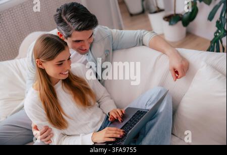 Affectionate young couple browsing a laptop together while sitting on a sofa in a bright modern living room Stock Photo