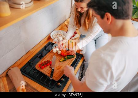 Overhead view of a young couple preparing fresh tomatoes, courgette, mushrooms and bell pepper on a wooden chopping board at home Stock Photo
