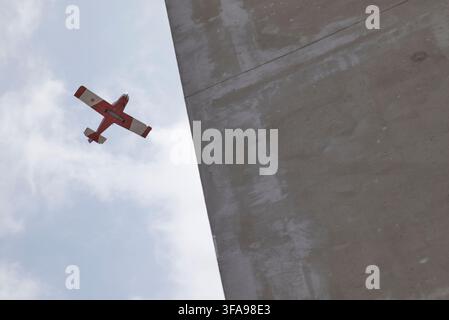 Small red airplane flying through a cloudy sky next to a tall brutalist concrete structure Stock Photo