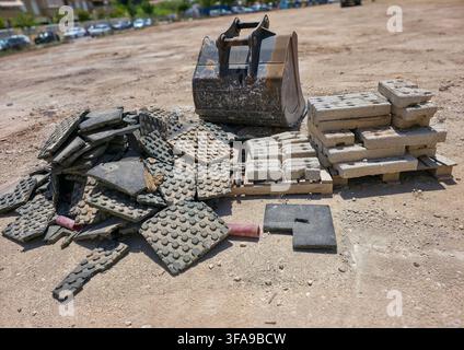 An excavator bucket rests on dirt at a construction site. Nearby, there are piles of discarded rubber tactile paving tiles and stacked concrete blocks Stock Photo