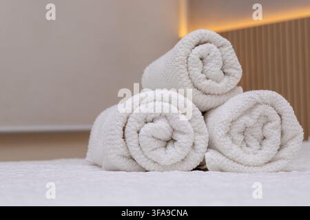 Three clean, fluffy white bath towels neatly rolled and stacked on a bed surface against a soft-lit modern bedroom interior background, representing h Stock Photo