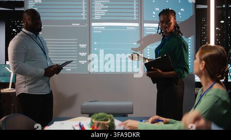 African american manager presenting monthly financial situation during night time meeting, reviewing resources available for a new project. Shareholders discuss possibilities. Camera A. Stock Photo