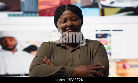 Portrait of cheerful woman celebrating freedom of unlimited video content in metaverse. Happy person stands with crossed arms in front of giant digital wall with online video channels, camera B Stock Photo