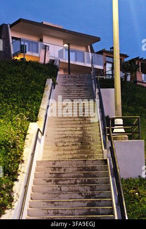 Staircase leads upward toward modern house on hillside. Green grass flanks concrete steps under evening sky. Metal railings guide ascent beside pole. Stock Photo