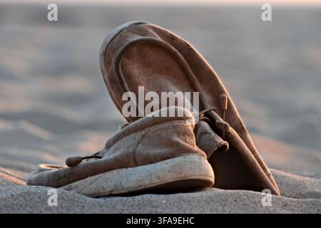 Worn leather moccasin shoes rest on soft sand. Warm golden light casts gentle shadows across the scene. Shoes appear well-used, hinting at past travel Stock Photo