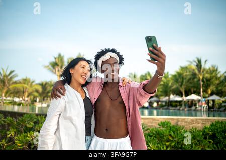 Trendy digital creator couple taking selfie by the beach, sharing travel content for social media Stock Photo