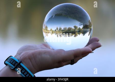 Close up of a female hand holding a crystal lensball reflecting a lake, wearing a smartwatch, with soft blurred  bokeh background Stock Photo