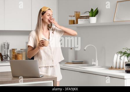 Morning routine and remote work lifestyle, sleepy woman yawning with coffee and laptop in a modern kitchen Stock Photo