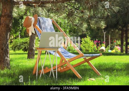 Remote work and digital nomad lifestyle, laptop on a small table next to a deck chair under a tree in a sunny park Stock Photo