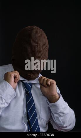 A faceless Asian male wearing a closed brown knitted balaclava stands against a deep black background while adjusting a crisp white shirt collar. The Stock Photo