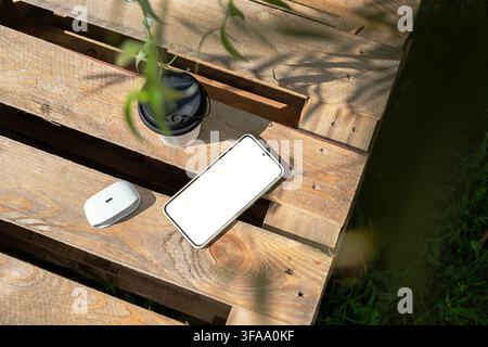 Smartphone, wireless earbuds and reusable coffee cup on rustic wooden table outdoors. Remote work, digital technology and productive lifestyle concept Stock Photo