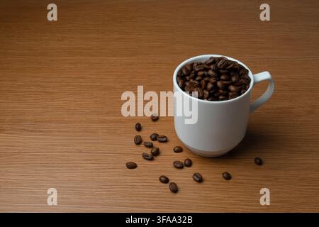 White mug filled with dark roasted coffee beans on a wooden tabletop with scattered beans. Copy space for text. Stock Photo