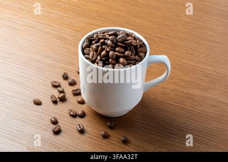 A white mug filled with roasted coffee beans on a wooden table with scattered beans. Copy space for text. Stock Photo