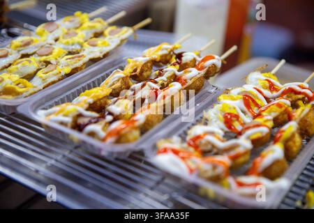 Rows of deep-fried street food skewers and fried quail egg sticks drizzled with mayonnaise and chili sauce sitting in plastic containers at an outdoor Stock Photo