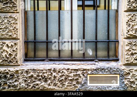 Close view of a frosted glass window protected by rusted iron bars in a heavily textured weathered facade, with an exterior ventilation grille below. Stock Photo