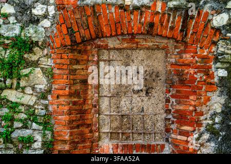 Blocked arched opening framed by weathered red bricks in an old stone wall, with concrete infill, iron bars and small plants growing between the mason Stock Photo