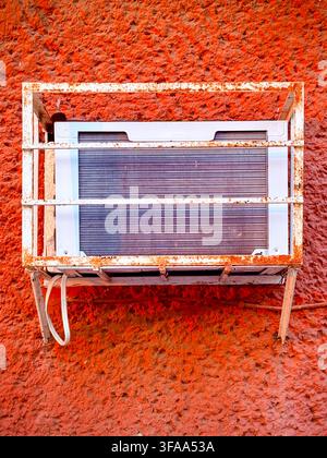 An old window air-conditioning unit in a protective metal cage on a bright wall in Mérida, Mexico, illustrating cooling demand and extreme heat Stock Photo