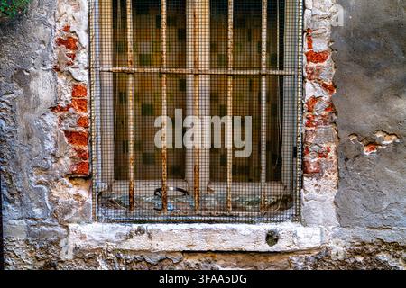 Old window protected by rusted metal bars and wire mesh, surrounded by exposed brick and worn plaster. Detailed architectural texture suitable for urb Stock Photo