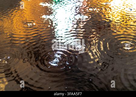 Raindrops produce concentric ripples and shifting reflections across a wet water surface, creating visible patterns of warm orange and bright blue lig Stock Photo