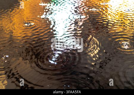 Close view of raindrops creating overlapping circular ripples across reflective water with vivid orange, white and blue light patterns. Stock Photo