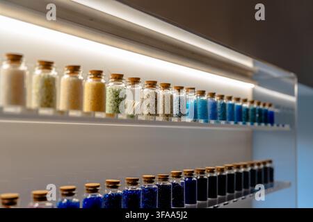 Rows of corked glass vials containing white, yellow and blue decorative beads displayed on transparent shelves beneath bright linear lighting. Stock Photo