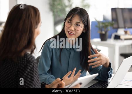 Two women working together at desk with laptop, using tablet, talking, collaborating on project, brainstorming, engaged in focused teamwork in office. Stock Photo