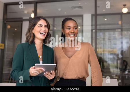 Two cheerful multinational businesswomen standing together in modern office, presenting ideas, engaged in friendly communication using tablet, laughin Stock Photo