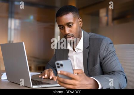 Focused African businessman sitting at office desk using laptop and smartphone, conducting digital communication, checking messages, reviewing busines Stock Photo