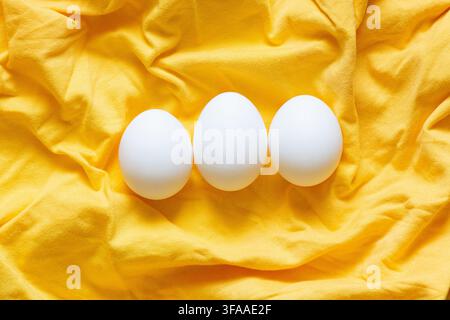 Three white chicken eggs on a bright yellow fabric, themed around food, natural product, breakfast, nutrition, and protein source Stock Photo