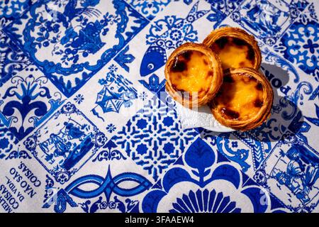 Three pastel de nata custard tarts bright pastry color in overhead top down view on blue and white tablecloth with traditional Portuguese azulejo tiles motifs pattern. Copyspace area. Translation: best things in life are not things. Stock Photo