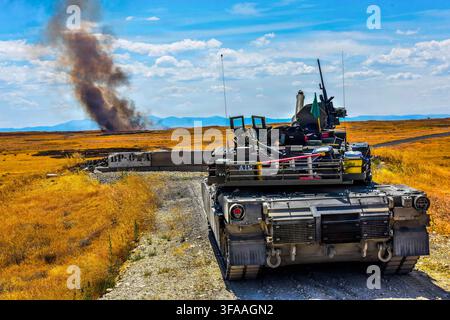 An M1A2 Abrams main battle tank sits stationary on the range as smoke rises from a grass fire in the background at Camp Orchard, Idaho, June 25, 2026, Stock Photo