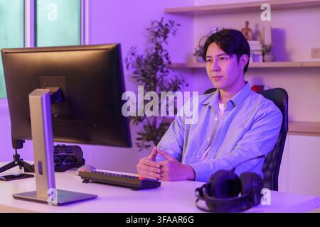 Asian man sitting at desk looking at computer monitor in neon purple lighted home office for remote work Stock Photo