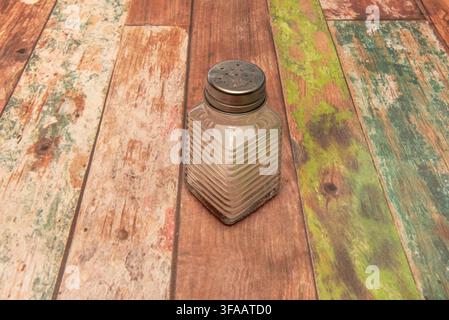 Empty glass salt shaker with perforated metal lid on colorful weathered wooden table surface. Stock Photo