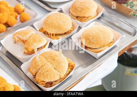 Row of crispy fried chicken burgers topped with melted cheese and sauces, packed in paper boxes on a metal tray. Stock Photo