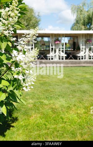 Beautiful white hydrangea flowers blooming in summer garden foreground. Luxury eco friendly resort with cozy outdoor restaurant terrace on green Stock Photo