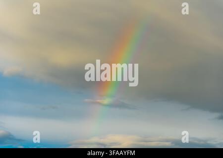 Vibrant multi-colored rainbow appears amidst dark, moody storm clouds and patches of blue sky, showcasing a beautiful natural phenomenon after rainfal Stock Photo