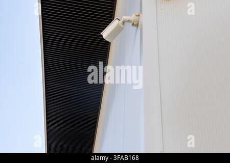 A white surveillance camera is attached to the exterior of a modern building with a ribbed white facade Stock Photo
