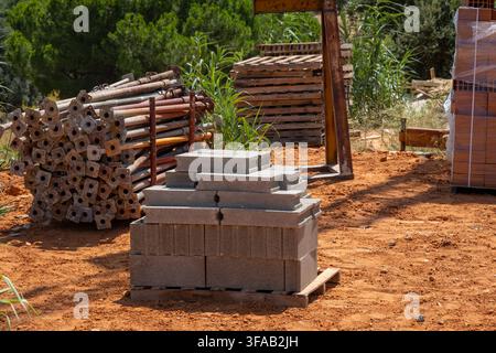 Various building materials including concrete blocks, scaffolding poles, and wooden pallets are stored on a construction site under bright sunlight Stock Photo