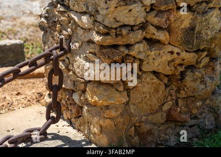 A thick, weathered chain is secured to a weathered, uneven stone structure under bright daylight Stock Photo