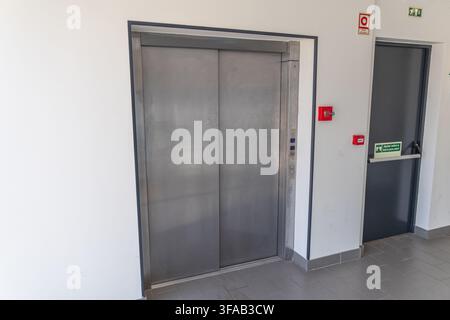 A contemporary elevator with brushed metal doors stands next to a dark gray emergency exit door with signage Stock Photo