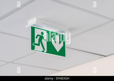 A modern green illuminated emergency exit sign is mounted on a suspended ceiling indicating the direction of escape Stock Photo