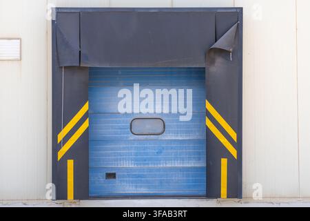 A closed blue roller shutter door at a warehouse loading bay, featuring yellow safety stripes and a dark grey protective frame Stock Photo