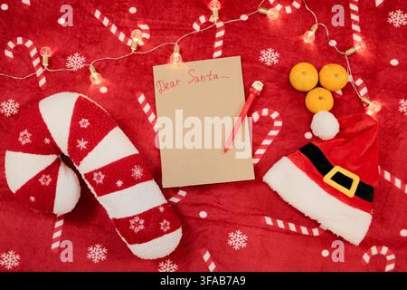 Festive flat lay with a blank Dear Santa letter, red pen, candy cane pillow, Santa hat, tangerines, and warm Christmas lights on a red holiday blanket Stock Photo