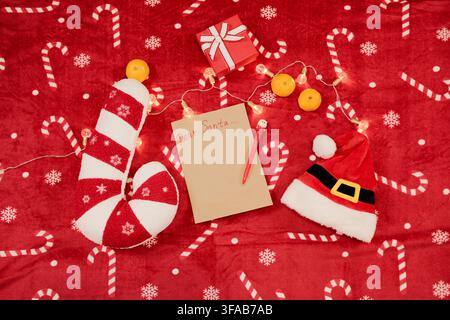 Festive flat lay with a blank Dear Santa letter, red pen, candy cane pillow, Santa hat, tangerines, and warm Christmas lights on a red holiday blanket Stock Photo