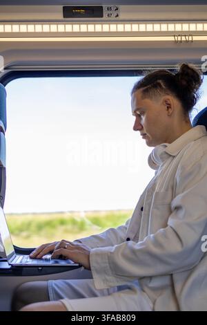 Mid adult digital nomad working on laptop near window inside passenger train carriage. Concept of flexible lifestyle remote employment on a trip and m Stock Photo