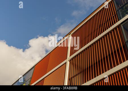 Modern building facade featuring repetitive orange vertical louvers and geometric patterns against a clear blue sky with clouds, urban design concepts Stock Photo