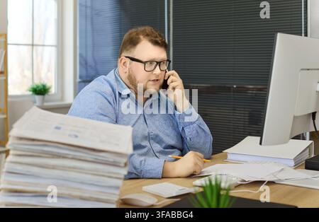 Focused overweight accountant on phone reviewing financial documents at computer Stock Photo
