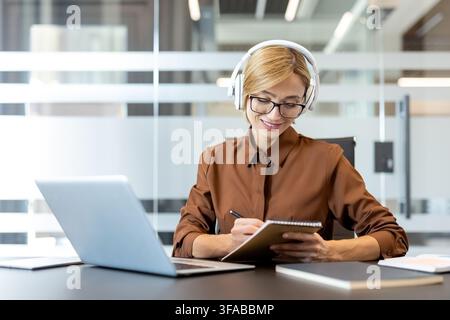 Focused businesswoman wearing eyeglasses and headphones, smiling while taking notes in a notebook next to a laptop during an online meeting or educational session in a modern office environment Stock Photo