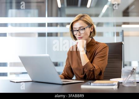 Businesswoman with short blonde hair and eyeglasses concentrating on her work, sitting at a desk and using a laptop in a contemporary corporate office environment Stock Photo